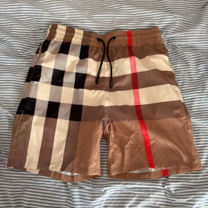 Rutiga Burberry BAD-Shorts - Snygga shorts med klassiskt Burberry-rutigt mönster i beige, brunt, svart och vitt med en röd linje. De har elastisk midja med svart snörning och är tillverkade i lätt polyester, perfekta för sommaren. Storlek S