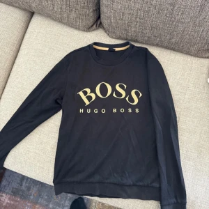 Hugo boss Sweatshirt - Stilren svart Hugo Boss sweatshirt med exklusiv guldfärgad Hugo Boss-logga på bröstet. Köpt från Zalando. Tyget och färgen är i mycket bra skick – den har inte tappat färg och ser fortfarande fräsch ut. Har lite slitage vid sista bilden men det är så lite så man märker inte ✔ Färg: Svart med guldig text ✔ Märke: Hugo Boss ✔ Skick: Mycket bra ✔ Köpt på Zalando 📦 Skickas inom 24h