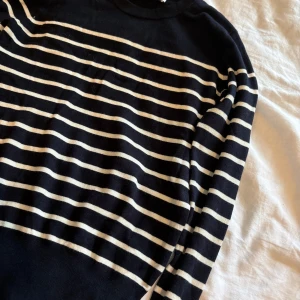 Randig stickad tröja från Zara - Mörkblå och vit randig stickad tröja från Zara med rund hals och långa ärmar. Snygga guldfärgade knappar vid ärmslutet ger en extra detalj. Perfekt för dig som gillar klassisk stil med en twist.