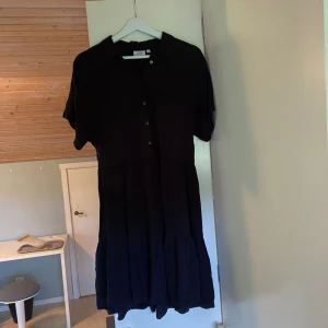 Svart kortklänning från KappAhl XS - Svart kortklänning från KappAhl i storlek XS. Klänningen har korta ärmar, knappar framtill och klassisk krage. Den har en luftig, volangig kjol och är tillverkad i ett mjukt tyg som känns skönt och luftigt.