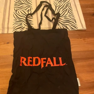 Redfall svart tygkasse med tryck - Svart shoppingväska i bomull med coolt rött tryck från Redfall. Ena sidan har texten 'REDFALL' och andra sidan har vampyrtänder och texten 'BITE BACK'. Perfekt för att bära böcker, laptop eller snacks.