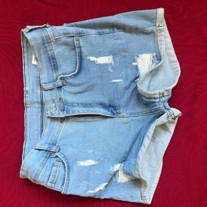 Ljusblå slitna jeansshorts - Säljer ett par ljusblå jeansshorts med slitna detaljer och råa kanter. Shortsen har klassiska fickor fram och bak samt bälteshällor. Perfekta för sommaren och ger en avslappnad vibe.
