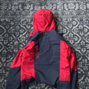 Röd och marinblå vindjacka från BOSS - Snygg vindjacka från BOSS i rött och marinblått med huva och dragkedja framtill. Jackan har två fickor med dragkedja och BOSS-logga tryckt på bröstet och ryggen. Tillverkad i lätt polyester, perfekt för blåsiga dagar.