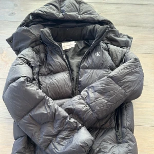 Svart pufferjacka från Calvin Klein Jeans - Svart pufferjacka från Calvin Klein Jeans med huva och dragkedja. Jackan har quiltad design, diskret CK-logga på bröstet och är vadderad för extra värme. Perfekt för kalla dagar och har en klassisk, stilren look.