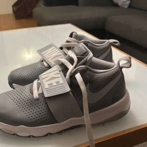 Grå Nike Team Hustle sneakers strl 40 - Säljer ett par grå Nike Team Hustle sneakers med vit sula och stora NIKE-detaljer på remmen. Skorna har snörning, perforerad tå och meshpartier för extra andningsförmåga. Perfekta för dig som gillar sportig stil och vill ha bekväma skor till vardagen.