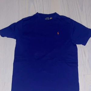 Blå t-shirt från Polo Ralph Lauren - Klassisk blå t-shirt från Polo Ralph Lauren med korta ärmar och rund hals. Ikoniska lilla broderade loggan i orange på bröstet. Skön och mjuk bomull som passar perfekt till jeans eller shorts.