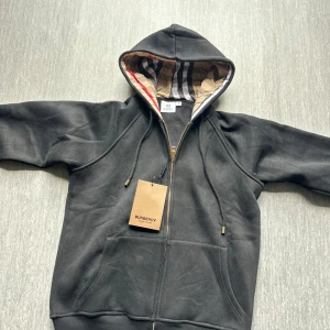 Svart hoodiejacka med mönstrad huva - Svart hoodiejacka från Nordic Spirit med dragkedja i guld och snörning i huvan. Insidan av huvan har ett klassiskt rutigt mönster i beige, rött och svart. Jackan har kängurufickor och är tillverkad i mjuk bomull, perfekt för en chill och stilren look.