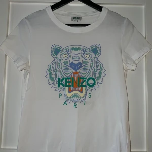 Vit Kenzo t-shirt med tryck - Säljer en vit t-shirt från Kenzo med ikoniskt tigertryck i grönt, blått och orange på bröstet. Klassisk rund halsringning och korta ärmar. T-shirten är i mjuk bomull och har en normal passform. I mycket bra skick och knappt använd. 