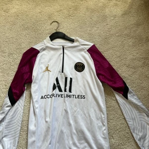 PSG x Jordan långärmad fotbollströja M - Snygg långärmad PSG fotbollströja från Jordan i vitt med vinröda och svarta detaljer på ärmarna. Tröjan har halv dragkedja framtill, broderad PSG-logga och Jordan-logga i guld. Tillverkad i lätt och ventilerande polyester, perfekt för träning eller match.