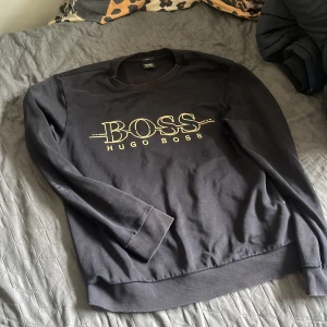 Svart BOSS sweatshirt från Hugo Boss M/S - Svart sweatshirt från Hugo Boss med stort BOSS-tryck i guld på bröstet. Klassisk rund halsringning och ribbade muddar vid ärmslut och nederkant. Mjuk bomullskänsla och långärmad modell, perfekt för en clean och stilren look.