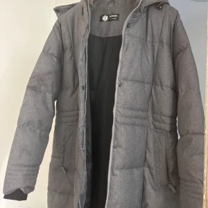 Grå pufferjacka från Tribute - Säljer en grå pufferjacka från Tribute med huva och dragkedja. Jackan är quiltad och har långa ärmar samt praktiska fickor på sidorna. Perfekt för kalla dagar och har en stilren look som funkar till det mesta.