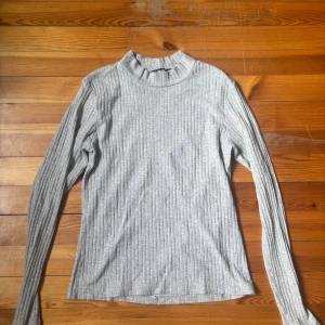 Grå ribbad långärmad tröja - Säljer en ljusgrå ribbad långärmad tröja från H&M med högre hals. Tröjan är i mjuk bomullsblandning och har en slim passform som sitter snyggt på kroppen. Perfekt till lager-på-lager och enkel att matcha med jeans eller kjol. Priset +frakt
