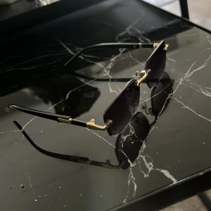 Svarta rektangulära solglasögon med gulddetaljer - Snygga solglasögon med rektangulära svarta linser och tunna svarta skalmar med gulddetaljer. Glasögonen har en minimalistisk design med metallinslag vid gångjärnen och näsbryggan. Perfekt accessoar för en cool och modern look.