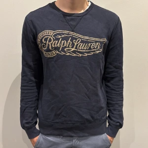 Mörkblå sweatshirt från Ralph Lauren - Snygg mörkblå sweatshirt från denim and supply Ralph Lauren med stort vintageinspirerat tryck i beige över bröstet. Tröjan har rund halsringning och långa ärma. Perfekt för en avslappnad och stilren look. Den är i storlek m. Skriv om ni har fler frågor