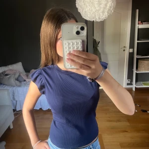 Mörkblå ribbad topp med volangärm - Säljer en mörkblå ribbad topp med korta volangärmar och rund hals. Toppen är figurnära och har en enkel men snygg design som passar perfekt till jeans. Materialet är stretchigt och bekvämt. Lite oversized för mig som är en XS passar S!! Kommer ej ihåg vart jag köpt eller beställt denna från.