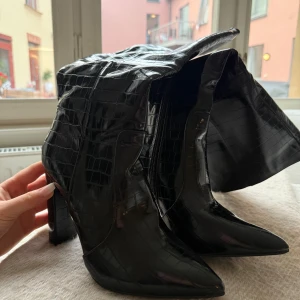 Svarta höga boots med krokopräglat mönster - Säljer ett par svarta boots med högt skaft och chunky klack. Skorna har spetsig tå och är gjorda i glansigt syntetmaterial med krokopräglat mönster. Dragkedja på insidan för enkel påtagning. Perfekta för att lyfta vilken outfit som helst. Helt nya och oanvända. Storlek 40