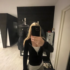 Svart velour hoodie från Juicy Couture - Svart hoodie från Juicy Couture i mjuk velour med dragkedja framtill och huva. Klassisk logga broderad på bröstet och känguruficka. Snygg och bekväm passform med långa ärmar, perfekt för chill dagar.