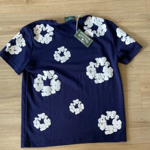 Blommig blå t-shirt från Denim Tears - Snygg mörkblå t-shirt från Denim Tears med vita blommiga tryck över hela plagget. T-shirten har klassisk rund hals och korta ärmar. Materialet är mjuk bomull som känns skönt mot huden. Perfekt för dig som gillar streetwear med unik design.