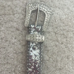 Glittrigt silverbälte med nitar - Säljer ett smalt bälte i silver med glittrig yta och dekorativa nitar längs kanterna. Spännet är prytt med strass och har en unik form. Bältet har stora metallhål och passar perfekt för att ge din outfit extra bling.