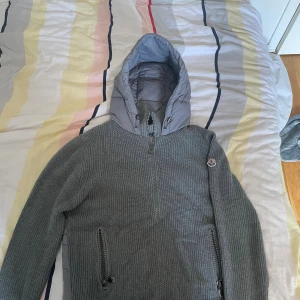 Grå Moncler cardigan - Snygg och stilren cardigan från Moncler i grå färg. Framsidan och ärmarna är stickade i mjukt material, medan rygg och huva är i vadderad nylon för extra värme och komfort. Jackan har dragkedja fram, två fickor med dragkedja och justerbar nederkant. Moncler-loggan sitter på ärmen. 	•	Färg: Grå 	•	Storlek: [M/L]