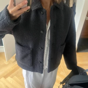 Mörkgrå teddyjacka från Uniqlo XS - Säljer en mörkgrå teddyjacka från Uniqlo i storlek XS. Jackan har en boxig, croppad passform med stora fickor framtill och knappar hela vägen. Materialet är mjuk och fluffig polyester som ger en cool och avslappnad vibe.