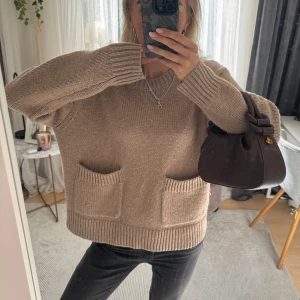 Beige stickad tröja med fickor - Mysig beige stickad tröja med två stora fickor framtill och breda ribbade muddar vid ärmslut och nederkant. Tröjan har en avslappnad, loose passform och är långärmad. Perfekt för lager-på-lager och chill dagar.
