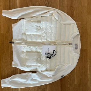 Vit Moncler cardigan jacka med dragkedja - Säljer en snygg vit cardigan jacka från Moncler med quiltad framsida och två fickor med knappstängning. Jackan har långärmade stickade ärmar och ribbade muddar. Dragkedja framtill och Moncler-logga på ena ärmen. Perfekt för dig som gillar stilren design.