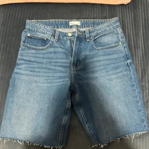 Blå jeansshorts med rå kant - Säljer ett par klassiska blå jeansshorts från Gina tricot med rå, fransig kant nertill. Modellen har fem fickor och normal passform. Perfekta för varma dagar och enkel att matcha med allt. Helt nya, finns dock ingen prislapp.