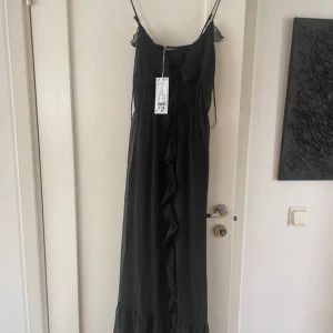 Ny Svart långklänning från Gina Tricot - Supersnygg svart långklänning från Gina Tricot i luftig chiffong med tunna axelband, volangdetaljer och snörning framtill. Klänningen har en romantisk vibe och faller snyggt längs kroppen – perfekt för dig som vill sticka ut.