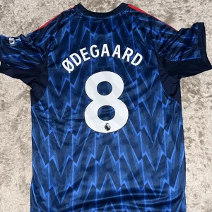 Arsenal Ødegaard bortatröja Adidas L - Säljer en Arsenal bortatröja med Ødegaard 8 på ryggen. Tröjan är mörkblå med ljusblå blixtmönster, röda Adidas-ränder på axlarna och klubbmärke samt sponsortryck framtill. Tillverkad i lätt och ventilerande material, perfekt för match eller träning.