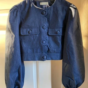 Mörkblå jeansjacka /blus från Vita Grace - Supersnygg mörkblå jeansjacka från Vita Grace med croppad passform, puffiga ärmar och dekorativa knappar framtill. Jackan har två fickor med lock på bröstet och vågig kant längs knappraden. Perfekt för dig som vill sticka ut med en unik denimlook. Jackan är oanvänd och köpt ny från Vita Grace.