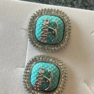 Turkosa örhängen med strass och Paris-logo - Säljer ett par fyrkantiga örhängen i turkos med quiltat mönster och silverfärgad Paris-logo. Kanterna är dekorerade med glittriga strasstenar som ger extra bling. Perfekt för dig som gillar statement-smycken och vill sticka ut.