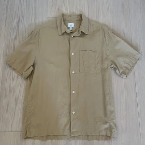 Beige kortärmad skjorta från H&M - Säljer en beige skjorta från H&M i relaxed fit. Skjortan har korta ärmar, klassisk krage, bröstficka och knäppning framtill. Tillverkad i ett svalt linnematerial som passar perfekt för varmare dagar.