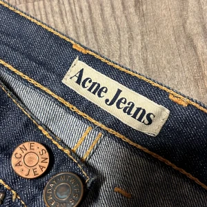 Acne studios jeans - Snygga mörkblå jeans från Acne Jeans i klassisk rak modell. Jeansen är tillverkade i 100% bomull och har orange kontrastsömmar, fem fickor och detaljerade knappar. Perfekta för dig som gillar stilrena och tidlösa jeans.