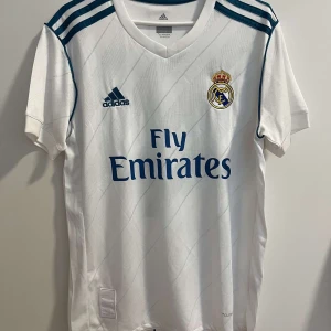 Real Madrid Sergio Ramos tröja - Vit Real Madrid matchtröja från Adidas med Sergio Ramos och nummer 4 på ryggen. Klassisk design med blå detaljer, klubbmärke broderat på bröstet och Fly Emirates-tryck. Tillverkad i lätt och ventilerande ClimaCool-material, perfekt för fotboll eller casual. Storlek S.