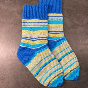 Randiga virkade strumpor i blått och gult 37/38 - Ett par handgjorda, virkade strumpor i glada färger. Strumporna har ett randigt mönster i blått, gult och grått, med en enfärgad blå mudd och tå. ﻿Bomull Fotlängd: 23 cm ﻿Skaft: 20 cm  Inga bilder när den sitter på mig! Katter finns i hemmet!