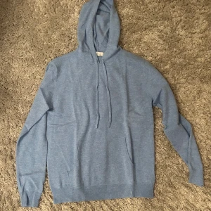 Blå Kashmir hoodie med huva och snörning - Mysig blå hoodie med huva och snörning framtill. Tröjan har långa ärmar, ribbade muddar och en klassisk känguruficka. Perfekt för dig som gillar en enkel och stilren look.