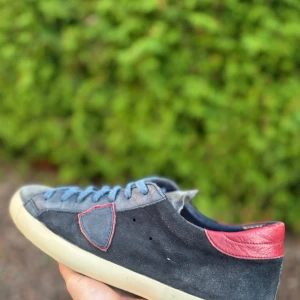 Philippe Model blå mockasneakers - Snygga sneakers från Philippe Model i blå mocka med grå detaljer och röd häl i skinn. Skorna har blå snörning, vit sula och den klassiska skölden på sidan. Perfekta för dig som gillar en sportig och stilren look.