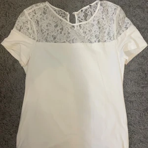 Vit blus med spetsdetaljer - Säljer en söt vit blus med korta ärmar och vacker spets upptill över axlar och bröst. Blusen har rund halsringning och en liten knapp i nacken. Perfekt för dig som gillar romantiska detaljer och en clean look. Står ingen storlek men skulle tippa på S/M☺️