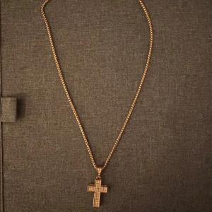 Guldfärgat halsband med korshänge - Stilrent halsband i guldfärg med ett korsformat hänge dekorerat med små glittrande stenar. Kedjan är tunn och elegant, perfekt för att ge din outfit en extra touch. Korset har en klassisk design som sticker ut och känns modern. pläterad guld.