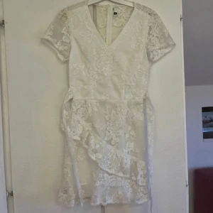 Vit spetsklänning med blommigt mönster - Superfin vit kortklänning med v-ringning och korta ärmar. Klänningen har ett vackert blommigt spetsmönster över hela och volangdetalj nertill. Perfekt för sommarens alla tillfällen! Materialet är spets och polyester för en luftig känsla. Klänningen är endast använd en gång men det finns några små fläckar, går säkert bort i tvätten. Bilderna visar fläckarna. 
