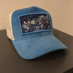 Blå och vit keps med Hv71-motiv - Säljer en blå och vit keps med mesh baktill och justerbar knäppning. Framsidan har ett stort Hv71-motiv med publik och flaggor i blågult. Kepsen har mjuk textil framtill och klassisk trucker-stil. Perfekt för dig som gillar sport och vill ha en stilren supporteraccessoar