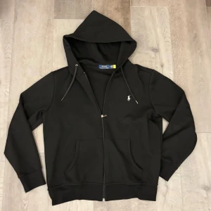 Svart hoodie från Polo Ralph Lauren - Svart hoodie från Polo Ralph Lauren med dragkedja, huva med snörning och klassisk broderad logga på bröstet. Tröjan har känguruficka framtill och är tillverkad i mjuk bomull. Perfekt för en chill och stilren look.