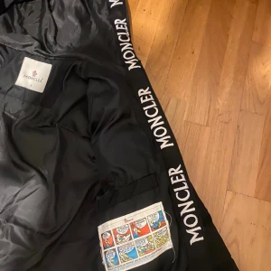 Svart Moncler dunjacka med huva - Svart dunjacka från Moncler med huva och dragkedja framtill. Jackan har stora vita Moncler-loggor längs insidan av huvan och öppningen. Två dragkedjeförsedda fickor och klassisk Moncler-märke på ärmen. Perfekt för kalla dagar.