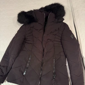 Svart pufferjacka från Amisu - Säljer en svart pufferjacka från Amisu med stor fluffig fuskpäls på huvan. Huvan är dessutom avtagbar. Jackan har quiltad design, två dragkedjefickor och snyggt emblem på ärmen. Perfekt för kalla dagar och riktigt skön att bära. Stängs med dragkedja hela vägen upp. Men har tidigare funnits ”skärp” till den men skärpet är borta och på sista bilden kan man se på sidan att det som höll upp skärpet har gått sönder men är en enkel fix.