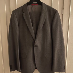 Grå kostym från Hugo Boss - Säljer en stilren grå kostym från Hugo Boss med diskret rutigt mönster. Kavajen har klassisk passform, två knappar framtill och fickor med lock. Byxorna har pressveck och hällor för bälte. Perfekt för dig som vill ha en snygg och tidlös look. Nyskick. Använd endast en gång. Original reservknapp finns i innerfickan. Köparen betalar frakt.