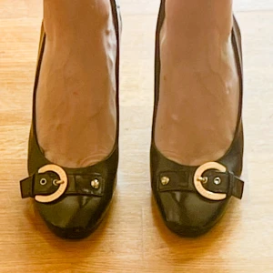 Louis Vuitton svarta pumps med gulddetalj - Snygga svarta pumps från Louis Vuitton i skinn med fyrkantig tå och bred klack. Skorna har en exklusiv guldfärgad metallspänne framtill med Louis Vuitton-logga och en elegant rem över vristen. Perfekta för dig som vill sticka ut med lyxiga detaljer. Fler bilder finnns. Sulan har förstärkts av skomakare.