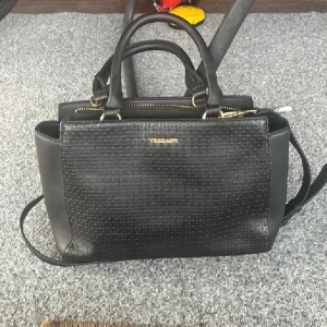 Svart handväska från Vizzano - Stilren svart handväska från Vizzano med präglat mönster på framsidan. Väskan har både handtag och axelrem, guldfärgade detaljer och dragkedja upptill. Tillverkad i skinnimitation och har flera fack för smart förvaring.