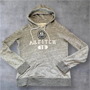 Grå hoodie från Abercrombie & Fitch - Snygg grå hoodie från Abercrombie & Fitch med broderad logga och text framtill. Tröjan har huva med snörning, magficka och långa ärmar. Perfekt för en chill och avslappnad stil.
