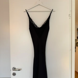 Svart långklänning med spetsdetalj - Supersnygg svart långklänning med smala axelband och djup v-ringning dekorerad med spets. Klänningen sitter lite lösare på kroppen men fortfarande figursydd, klänning är i siden och använd en gång💕